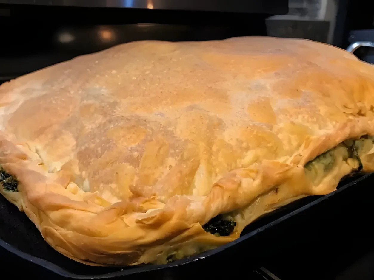 A golden layered byrek in a rectangular black cast-iron pan, with the spinach filling showing at the edges