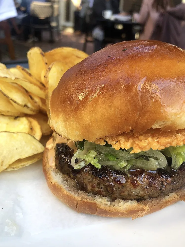 The Secret Burger at Alden & Harlow on a glossy brioche bun with shredded lettuce and a crispy cheese layer, next to potato chips