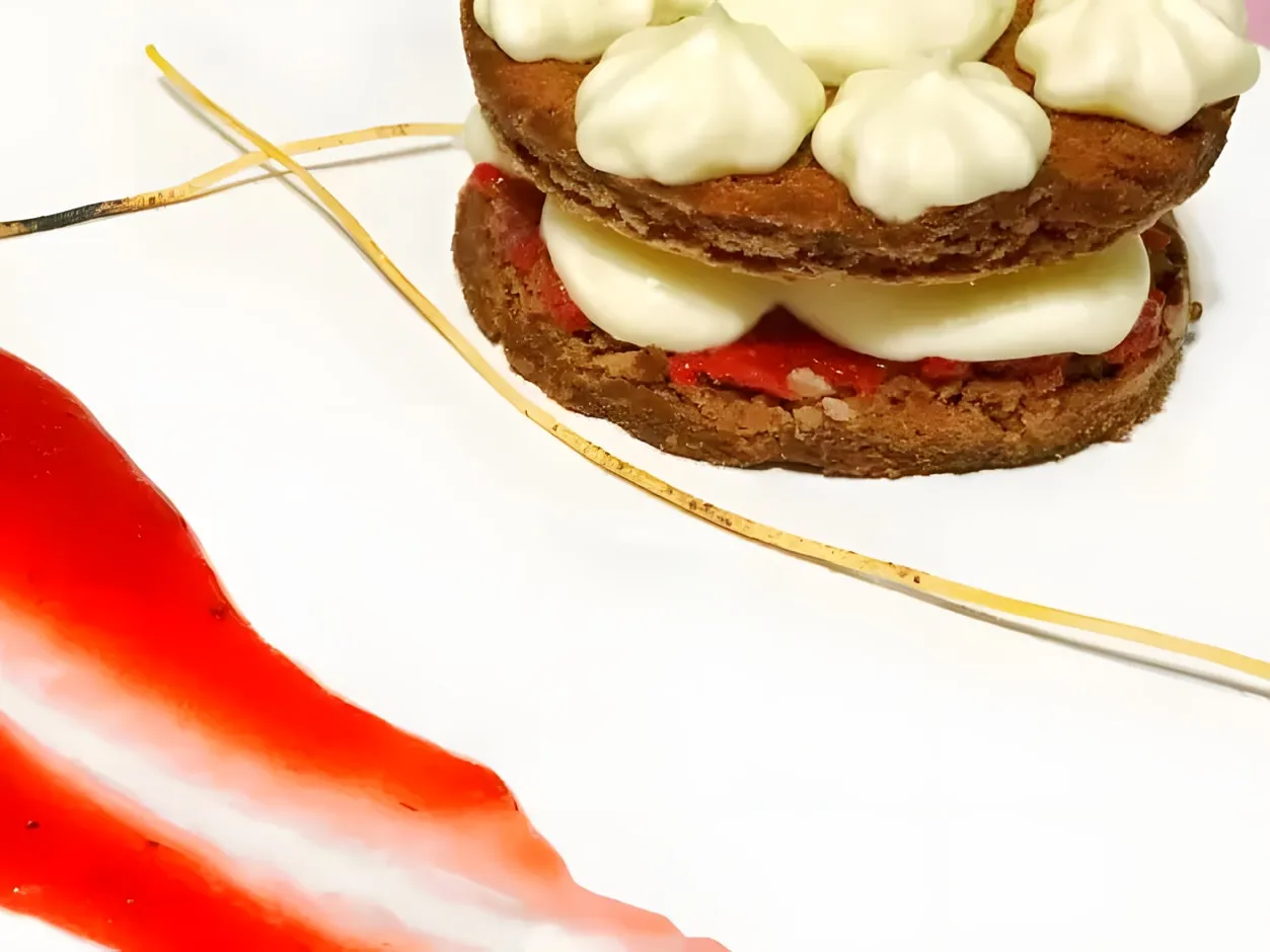 Two rounds of chocolate cake with strawberry pieces and then cream cheese frosting between them, more frosting piped on top, next to a swipe of strawberry sauce and thin golden strands on a white plate