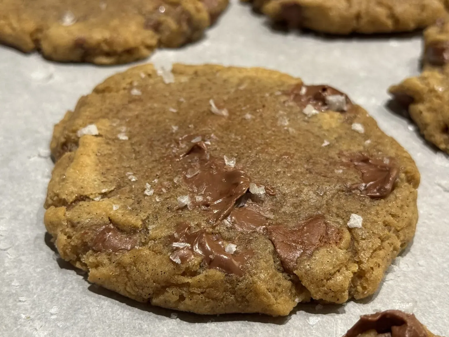 A chocolate chip cookie with melted chocolate and flakes of sea salt on top, on parchment paper