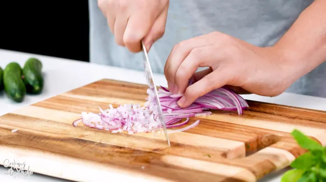 Hands chopping thin slices of red onion into small pieces on a wooden board, with small cucumbers and basil beside it