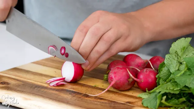 Hands slicing a radish paper-thin with a chef’s knife on a wooden board, next to a bunch of radishes with their leaves