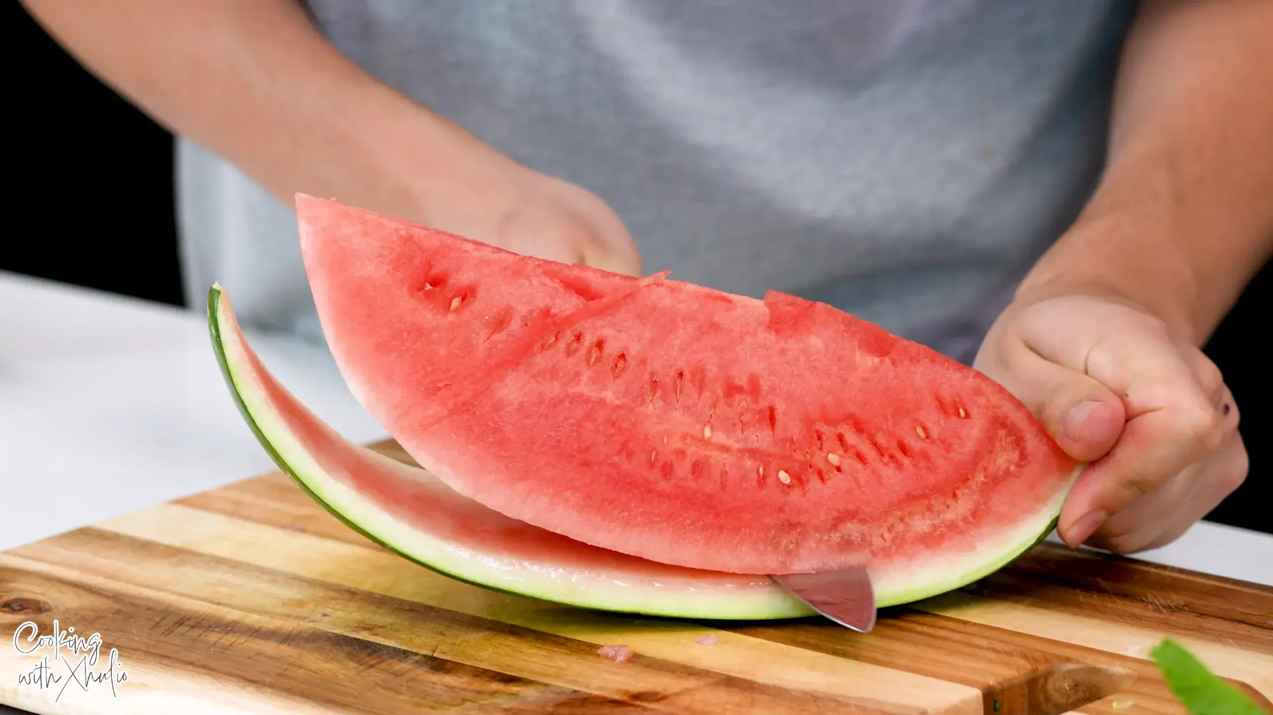 A watermelon wedge on a wooden board, with the knife cutting along the line where the red meets the white rind