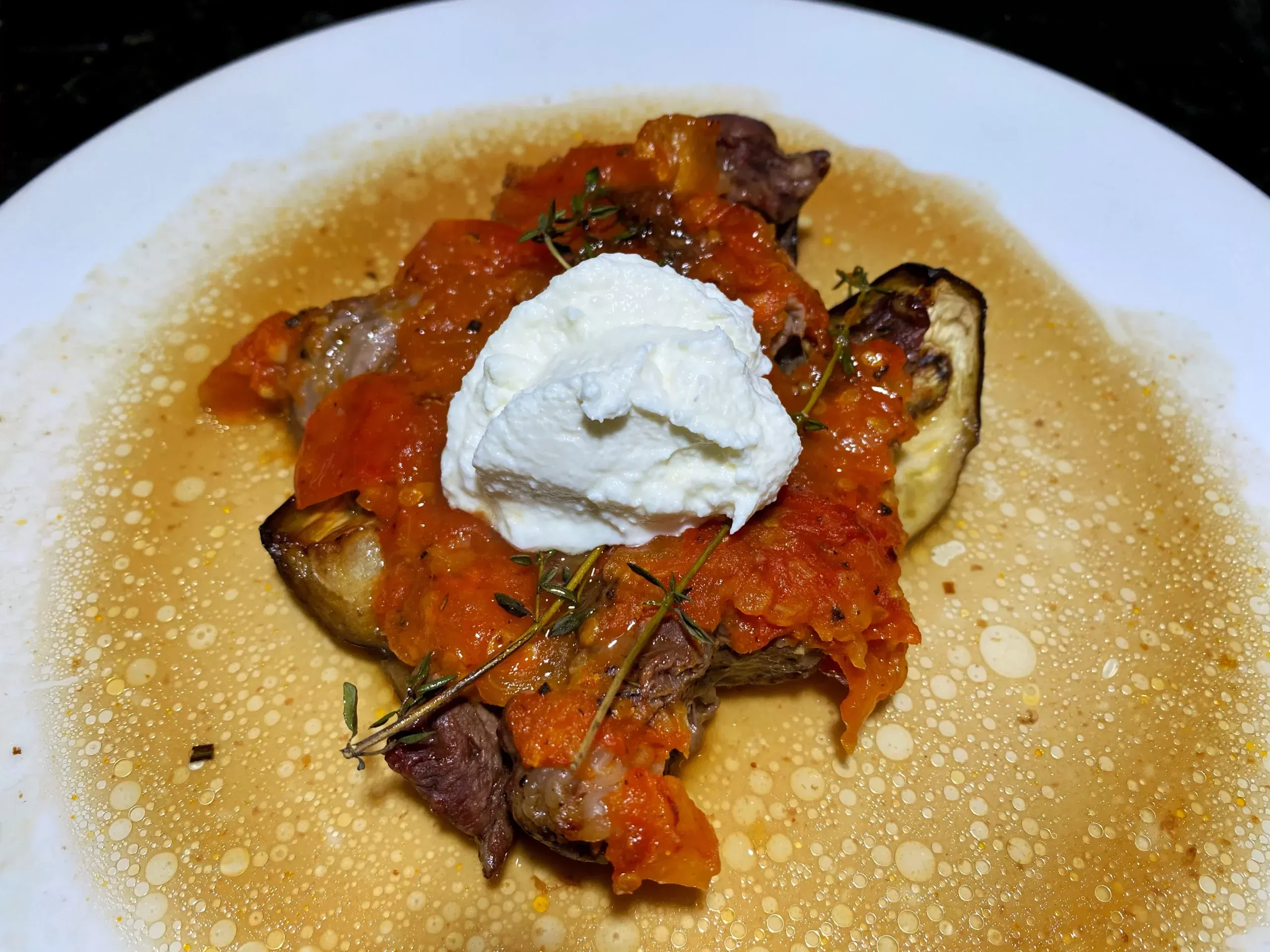 Baked eggplant under tomato sauce and pieces of beef, with a spoonful of soft white farmers cheese and sprigs of thyme on top, on a white plate