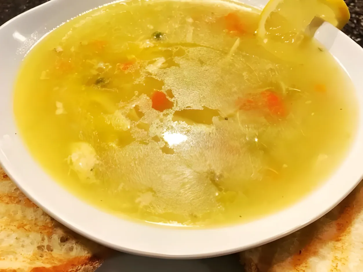 A white bowl of clear chicken noodle soup with carrots and shredded chicken, a slice of lemon on the rim and grilled bread on both sides