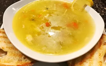 A white bowl of clear chicken noodle soup with carrots and shredded chicken, a slice of lemon on the rim and grilled bread on both sides