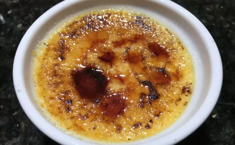 A crème brûlée in a white ramekin with a dark, torched sugar crust, on a black stone counter