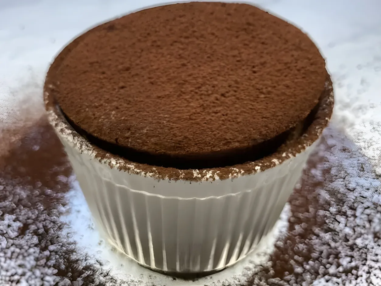 A risen chocolate souffle dusted with cocoa powder in a white fluted ramekin, with cocoa sprinkled on the counter around it