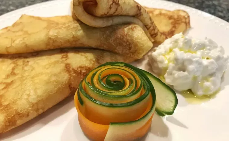 Folded golden crepes on a white plate with cottage cheese, a drizzle of oil and a rose of cucumber and carrot ribbons