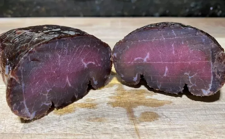 Homemade pastirma cut in half on a wooden board, deep red inside with a dark, glossy crust