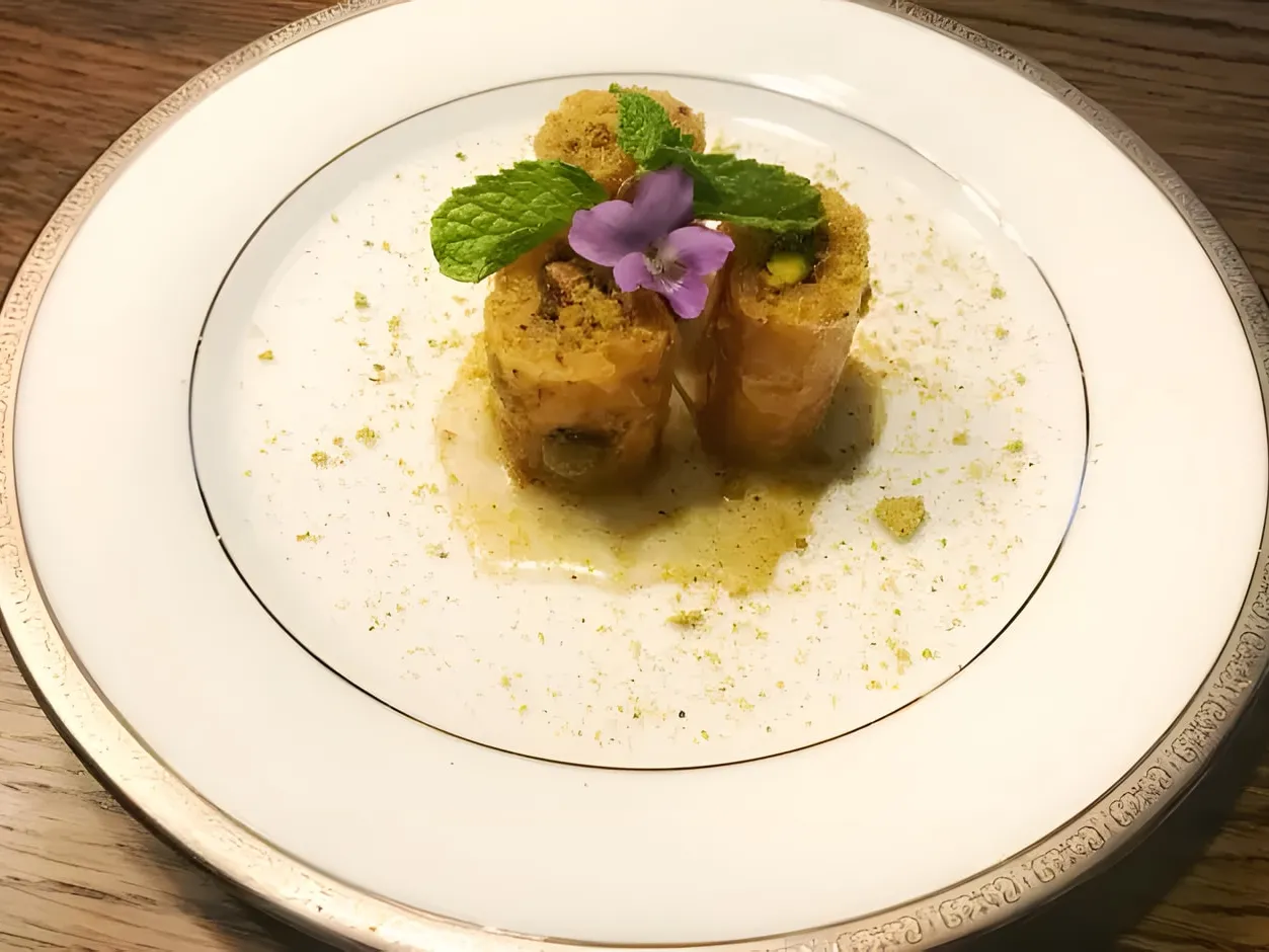 Three pieces of pistachio baklava fingers standing on a white plate, topped with mint leaves and a small purple flower, with crushed pistachios and syrup around them