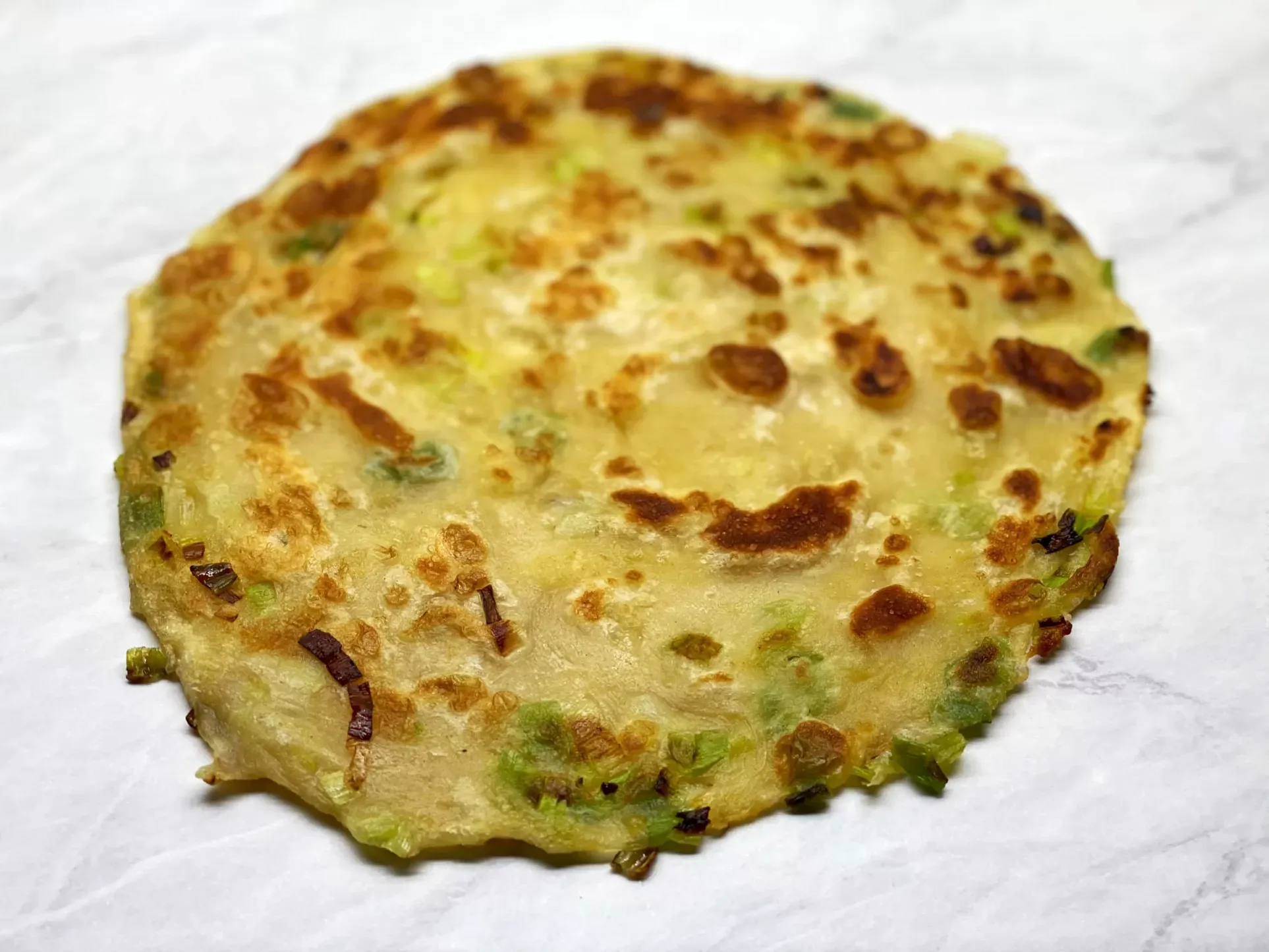 A golden scallion pancake with crispy brown spots and bits of green scallion, on a white marble counter