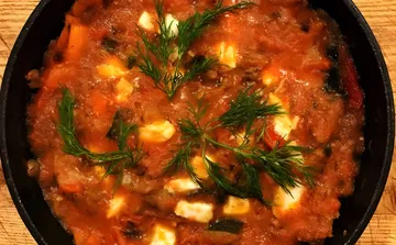 Turli in a black cast-iron pan, with peppers, zucchini and eggplant in a thick tomato sauce, cubes of mozzarella and sprigs of dill on top