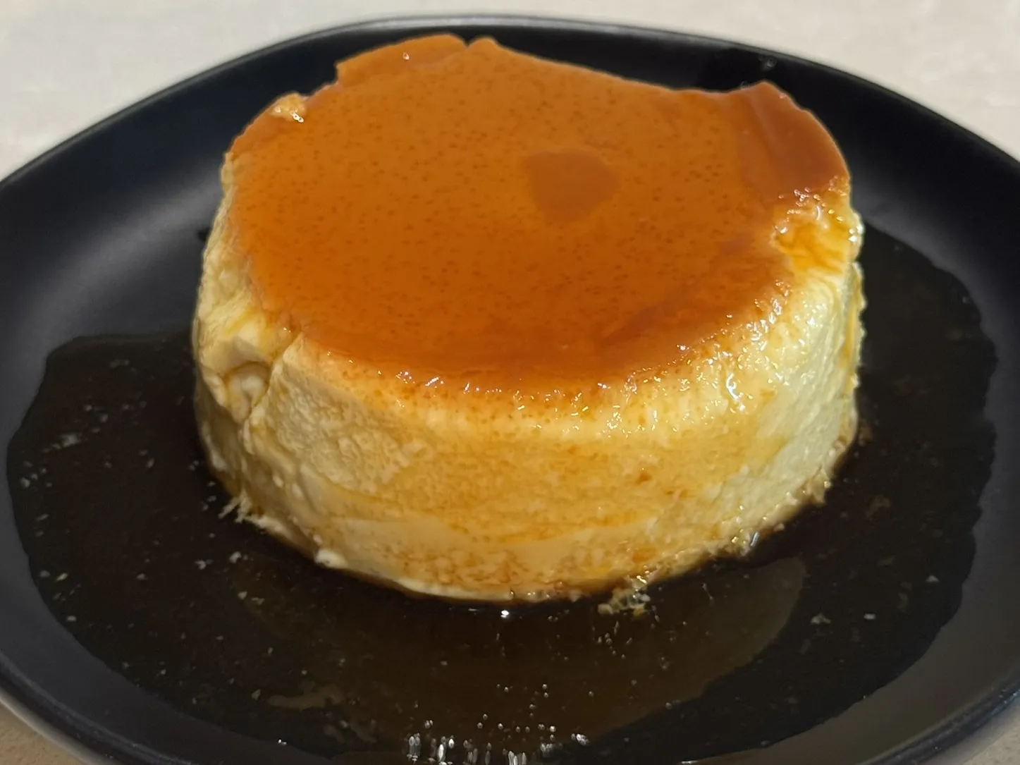 A crème caramel turned out on a black plate, with dark caramel sauce pooled around it