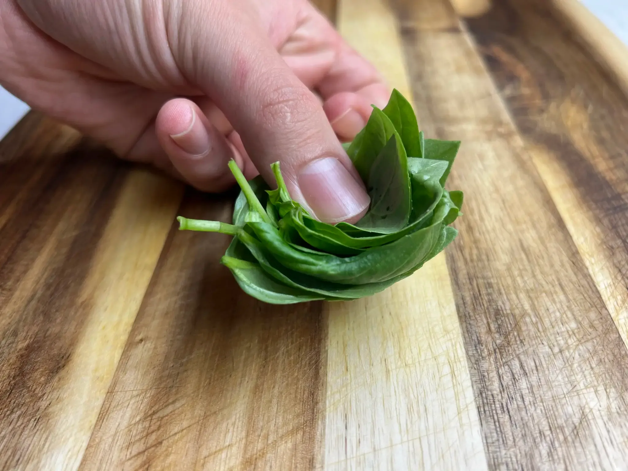 A hand holding a stack of fresh basil leaves nested into each other, on a wooden cutting board
