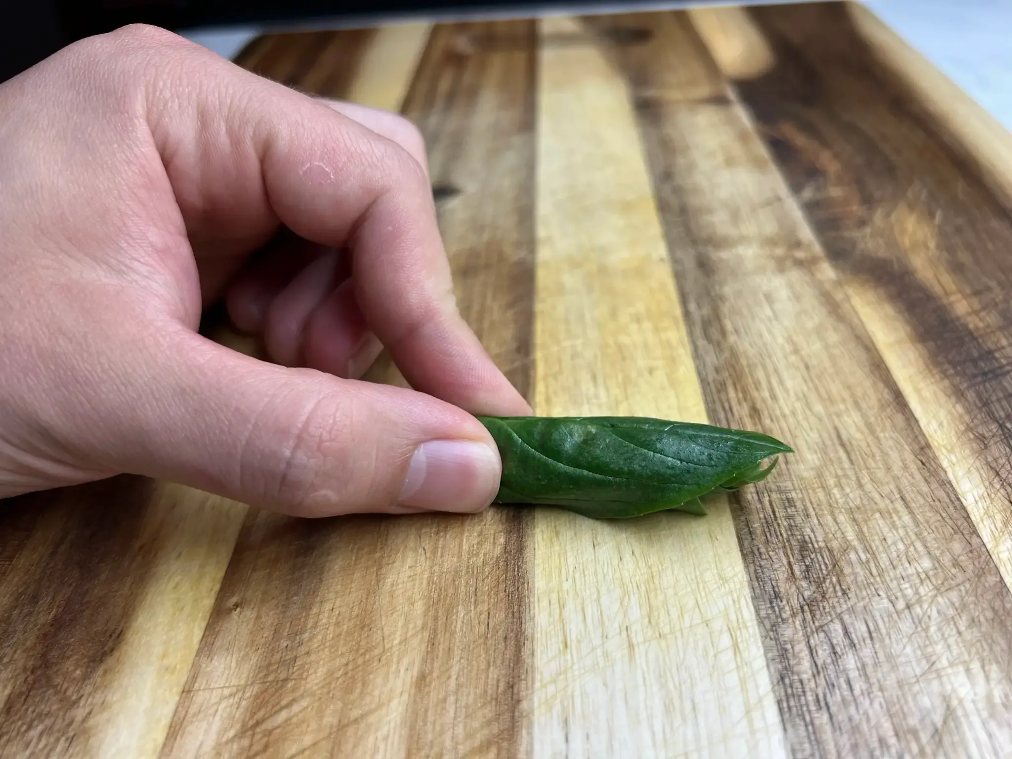 Basil leaves rolled tightly into a thin cigar shape, pinched between thumb and finger on a wooden cutting board