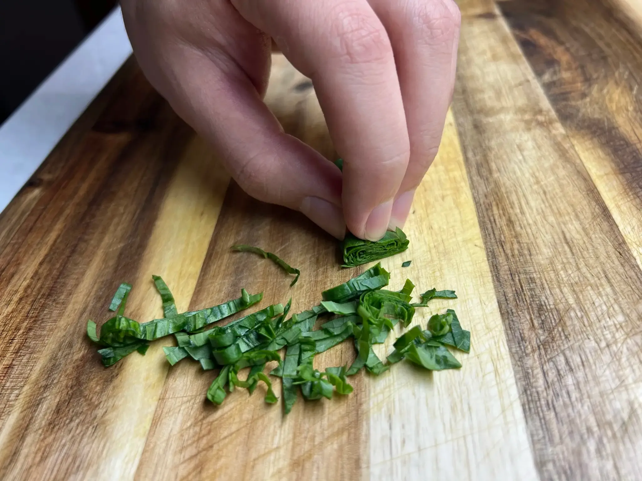 Fingertips holding the end of the basil roll, with freshly sliced thin strips of basil on the board in front