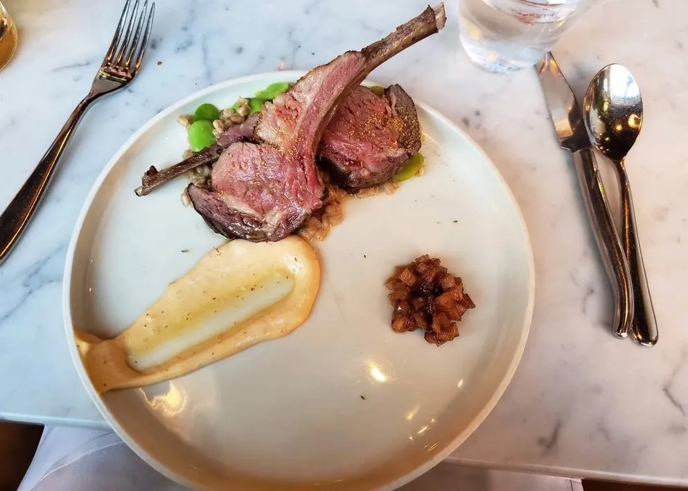 Two pink lamb chops on grains and fava beans, with a swoosh of creamy sauce and a spoonful of diced relish, on a marble table at Pammy’s