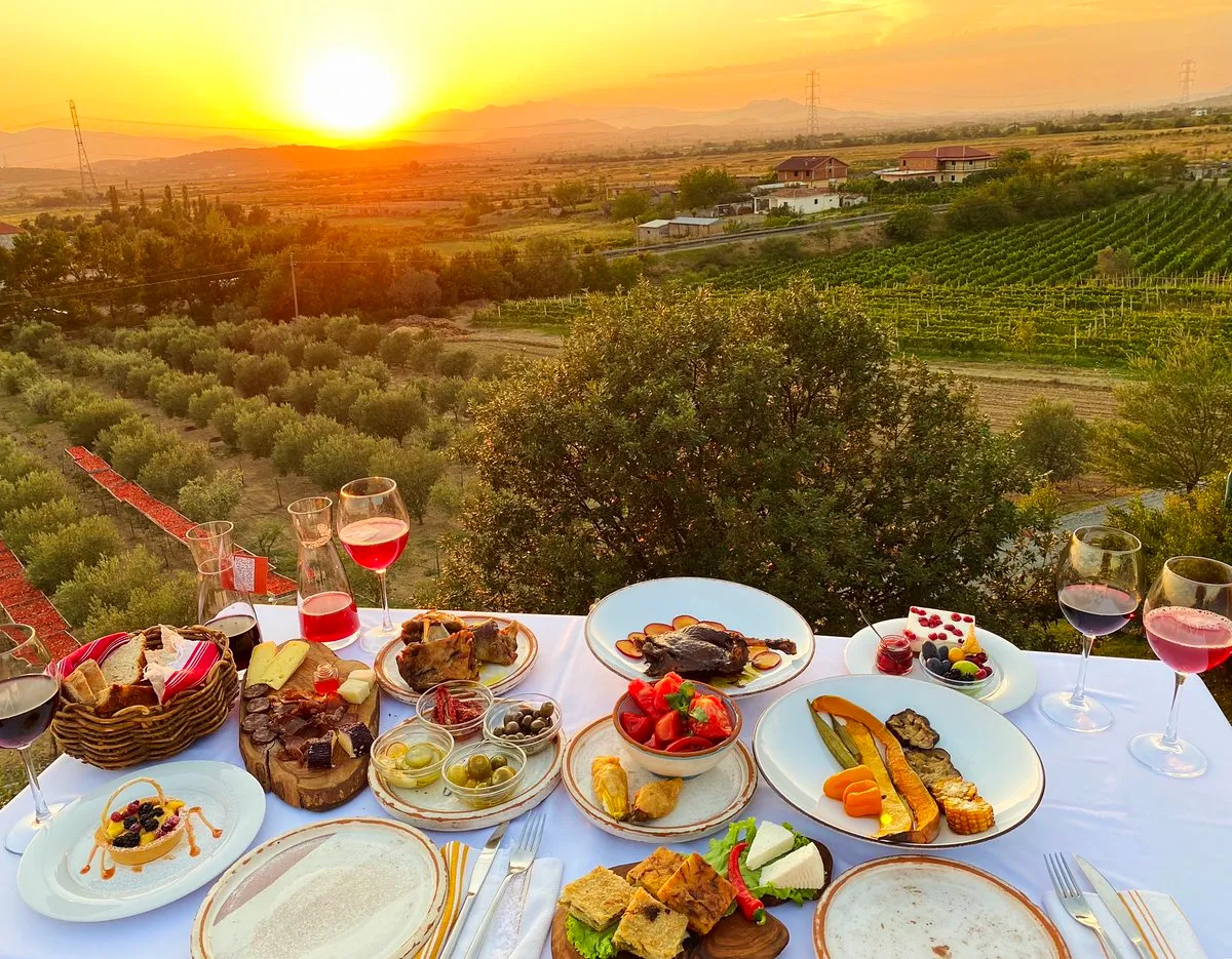 A table at Mrizi i Zanave full of bread, cheese, cured meats, olives, tomatoes, zucchini blossoms, grilled vegetables, desserts and wine, above olive groves and vineyards at sunset