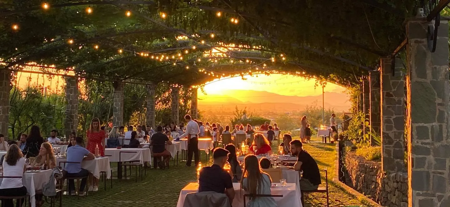 Diners at tables with white cloths under a vine-covered pergola strung with lights, as the sun sets over the hills at Mrizi i Zanave