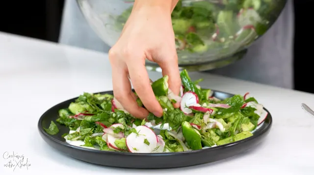 A hand placing the cucumber and radish salad from a glass bowl onto a black plate spread with Greek yogurt