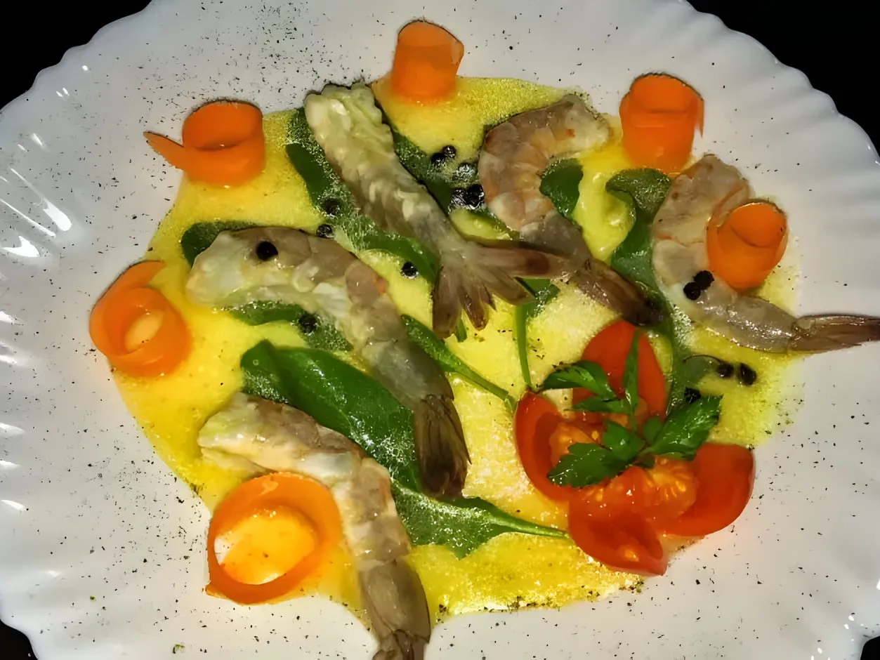 Raw shrimp crudo on a white scalloped plate, in a yellow lemon and olive oil sauce with arugula, rolled carrot strips, black peppercorns and a cherry tomato cut into wedges with a parsley sprig