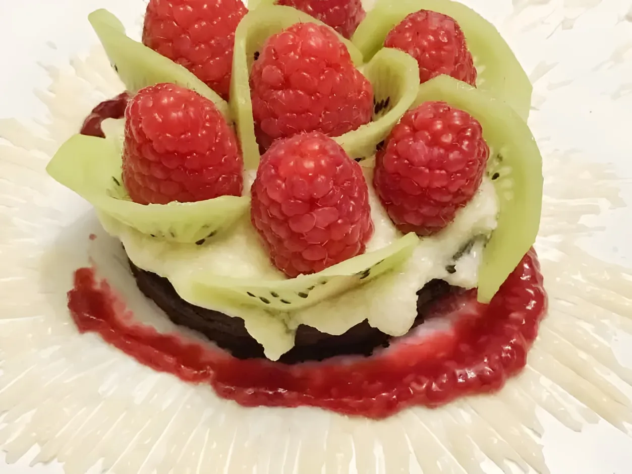 A round of chocolate brownie covered in vanilla frosting, with curved kiwi slices standing up between fresh raspberries, on a swipe of raspberry sauce and a white starburst painted on the plate