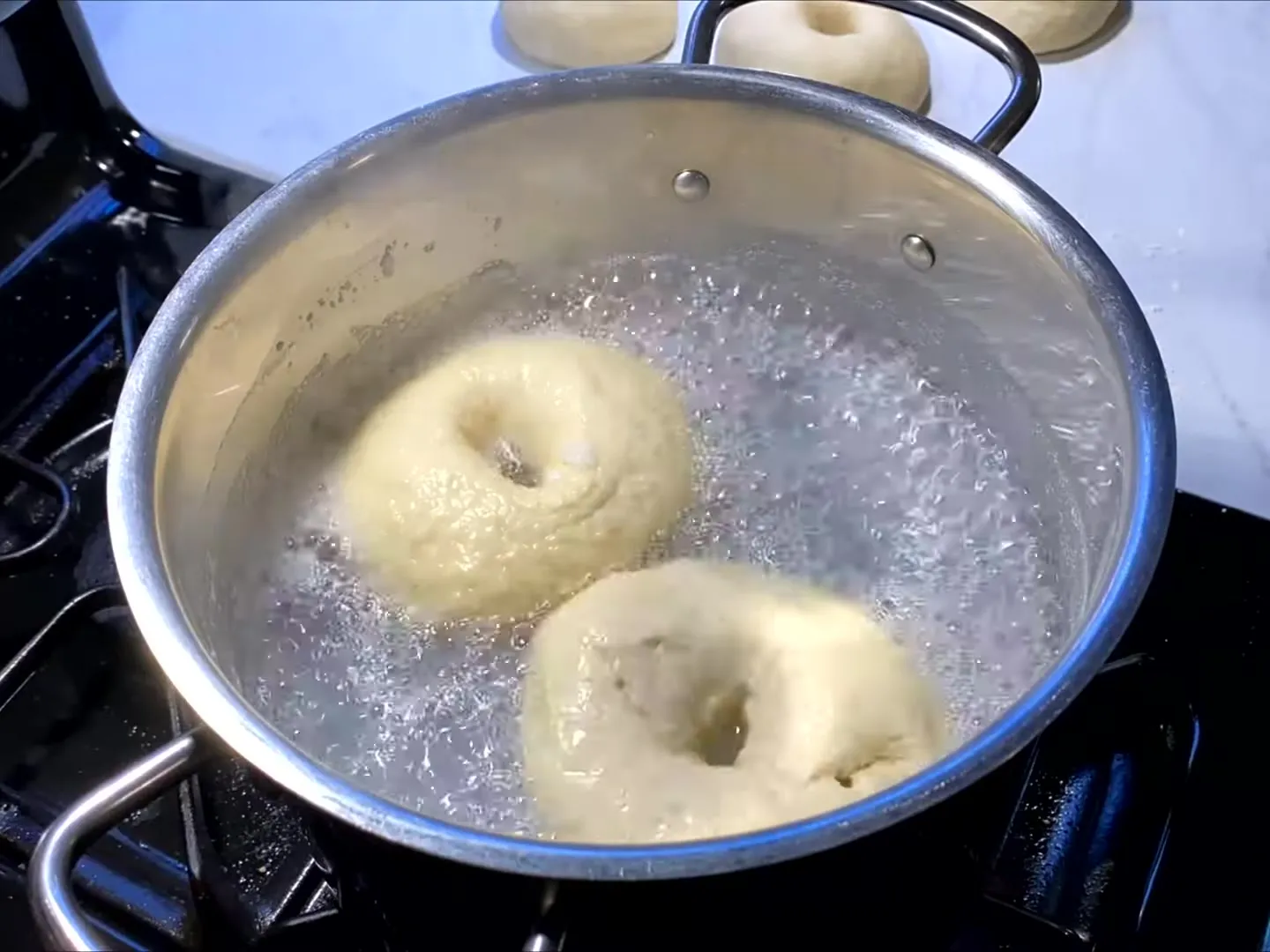 Two bagels boiling in a wide steel pot of bubbling water, with more shaped bagels waiting on the counter behind it