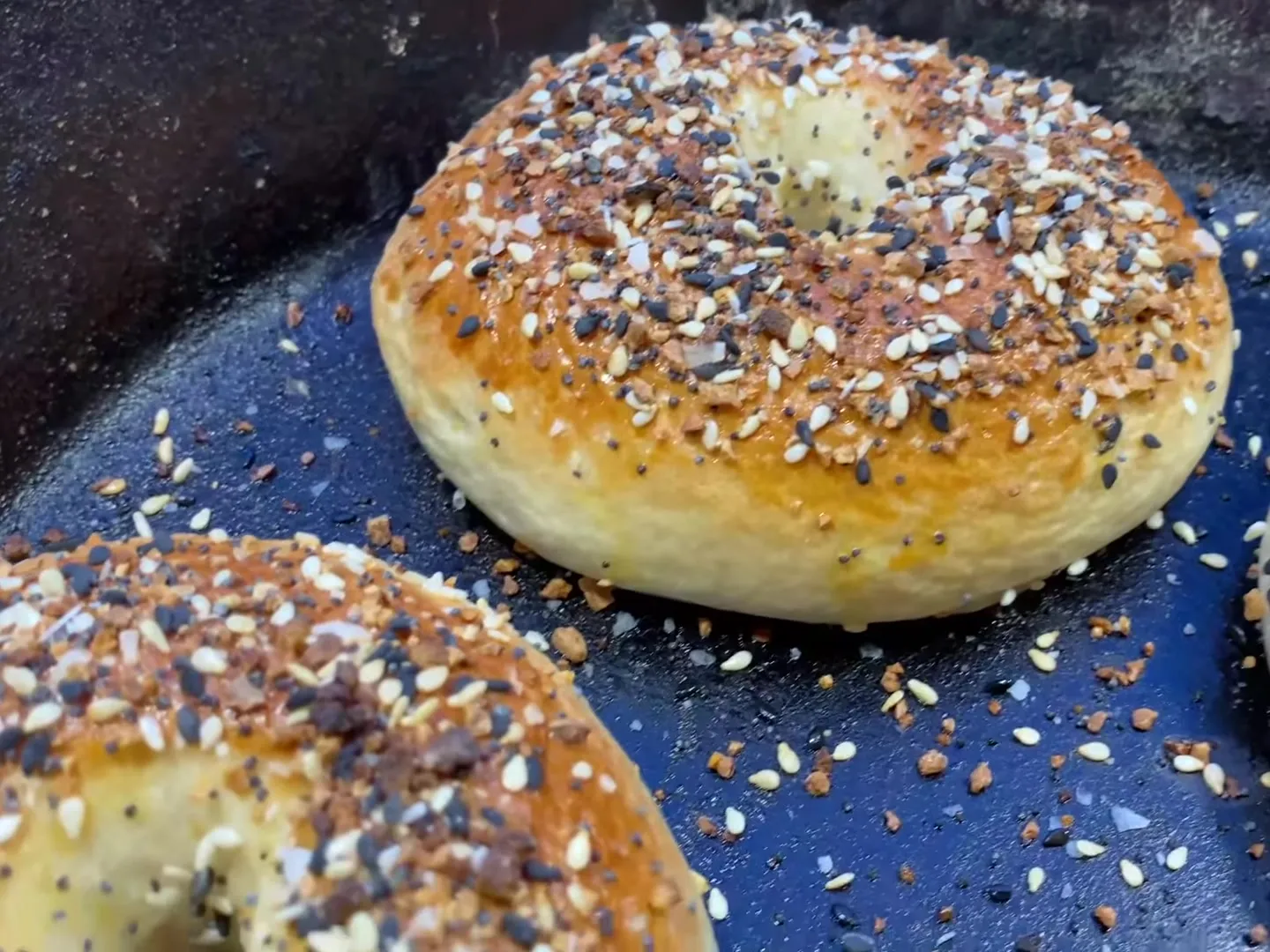A golden homemade everything bagel in a cast-iron skillet, with seeds scattered around it
