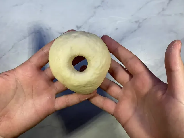 A shaped bagel held in two hands, with a round hole in the middle, over a marble counter