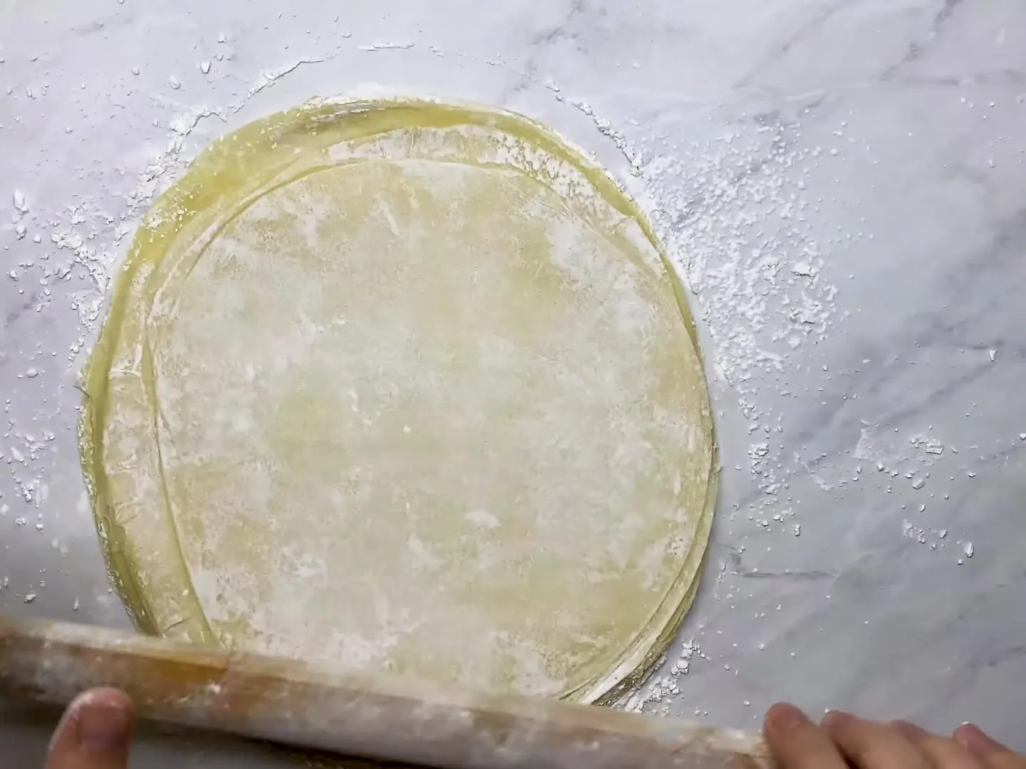 A stack of starch-dusted phyllo circles on a marble counter, being rolled out together with a long rolling pin