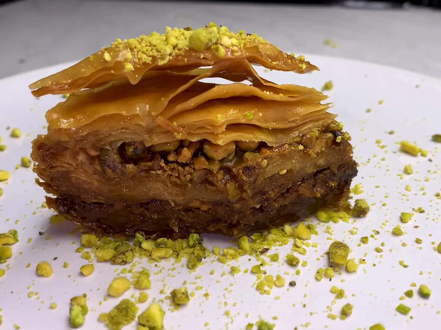 A piece of homemade baklava on a white plate, showing its thin golden layers and the pistachio filling, with crushed pistachios around it