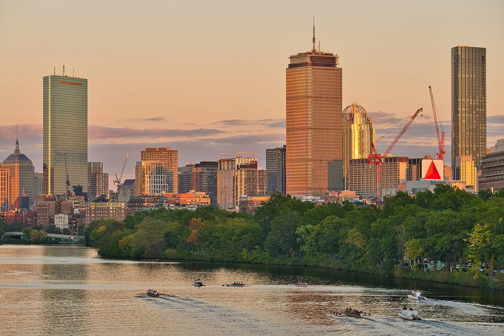 Boston's Back Bay skyline in golden evening light across the Charles River, with the Prudential Tower, the Citgo sign and rowing crews on the water