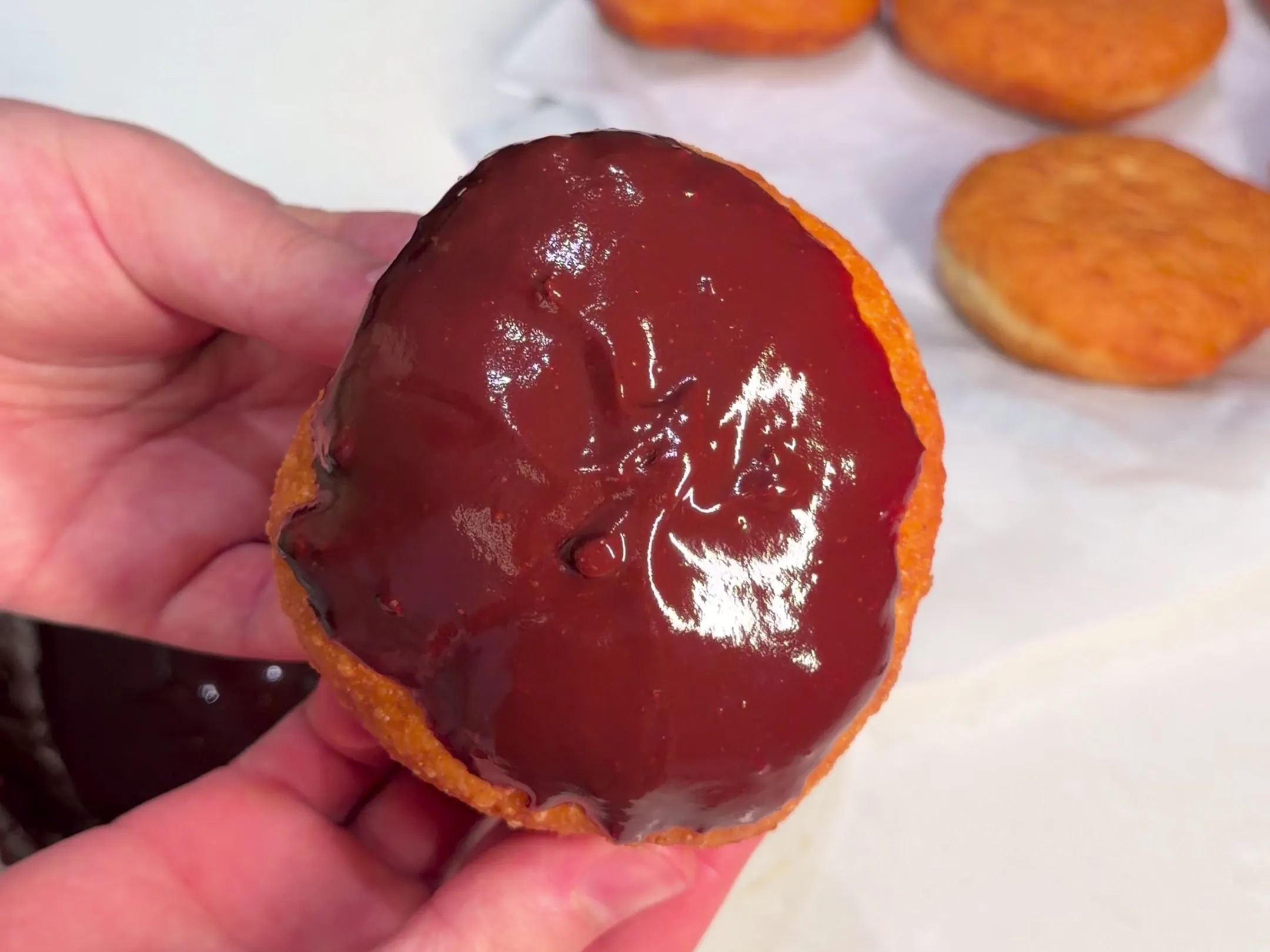 A fried donut held up with its top dipped in glossy melted chocolate, plain fried donuts on paper behind
