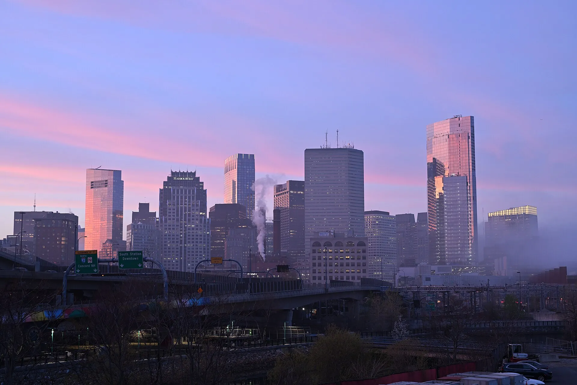 Downtown Boston's towers in pink sunset light and mist, seen from West Broadway in South Boston, behind the I-93 overpass and its green highway signs