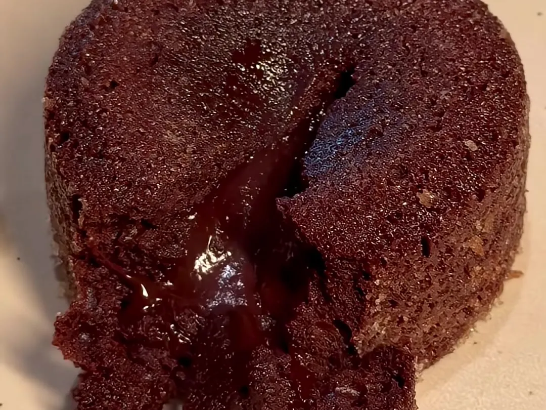 A chocolate lava cake turned out on a cream speckled plate, with a wedge cut from the front and the glossy molten chocolate center running out