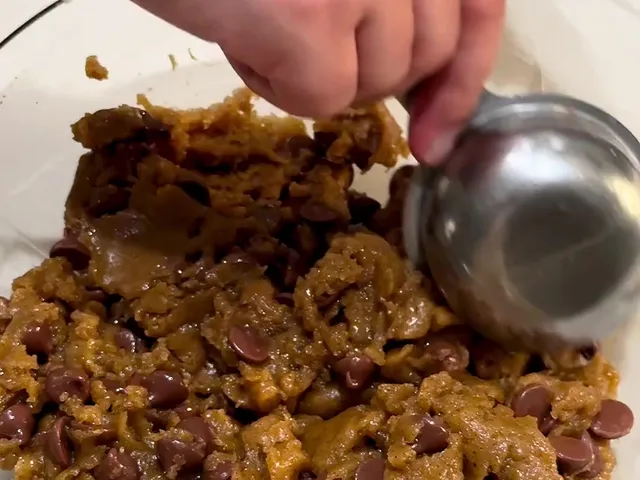 Chocolate chips mixed through the cookie dough in a glass bowl, with a metal measuring cup scooping out the first cookie