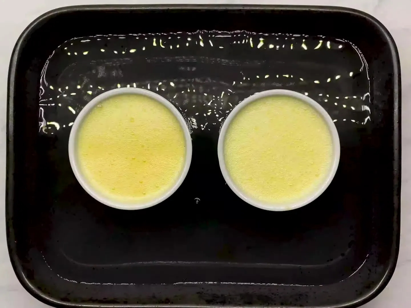 Two white ramekins of custard sitting in hot water in a black baking dish, ready for the oven