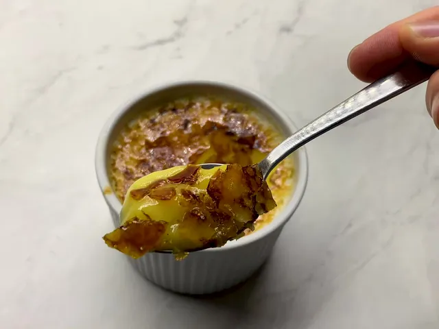 A spoonful of crème brûlée lifted from the ramekin, yellow custard with shards of the caramelized sugar crust on top