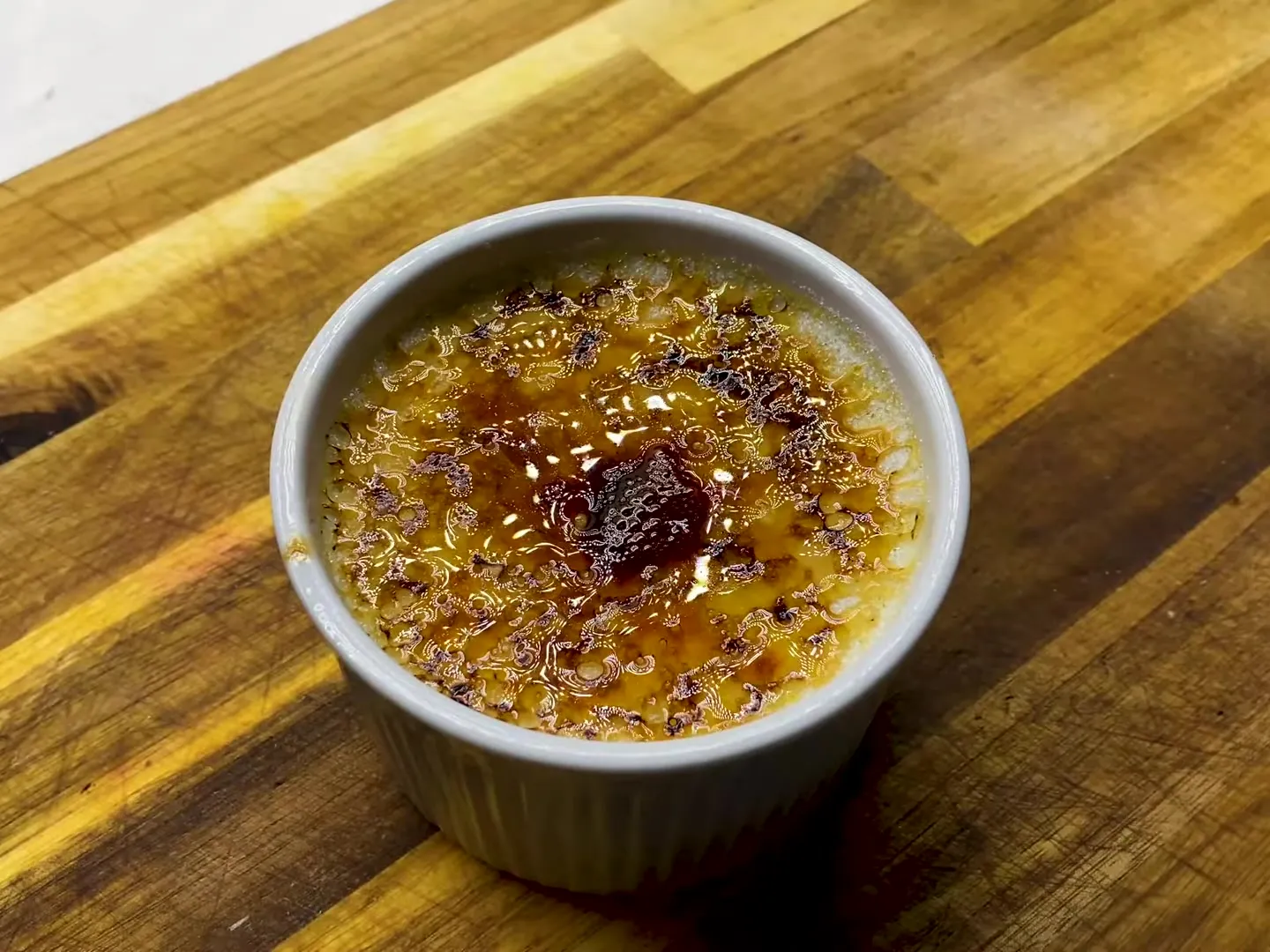 A crème brûlée in a white ramekin with a glossy, torched sugar crust, on a wooden board
