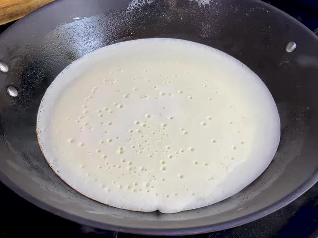 Crepe batter spread thin across a black pan, with small bubbles on top