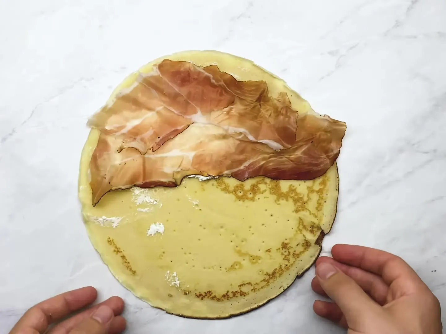 A crepe on a marble counter with cream cheese and a slice of prosciutto on one half, ready to fold