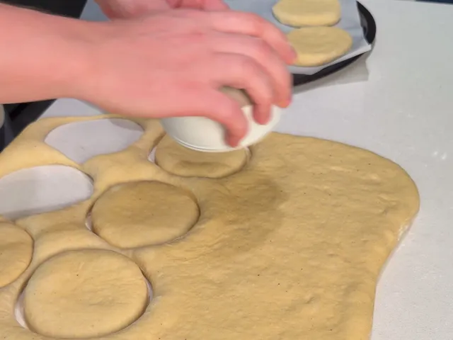 Cutting circles out of the dough sheet with an upside-down small white bowl, with cut rounds on a tray behind