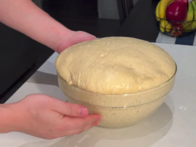 Hands holding a glass bowl of risen donut dough, domed above the rim
