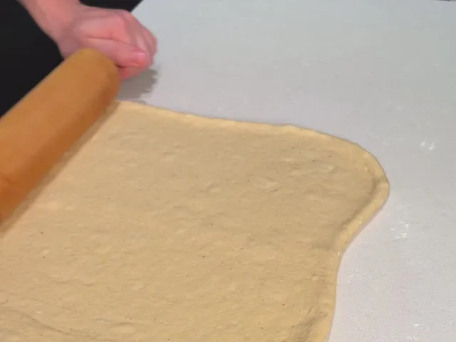 A rolling pin at the edge of an even sheet of donut dough on a floured white counter