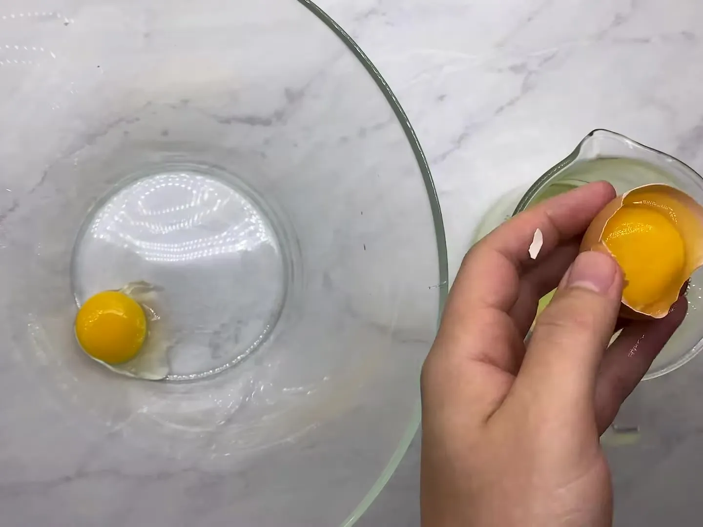 A hand holding an egg yolk in half of the shell over a measuring jug of egg whites, with one yolk already in a glass bowl
