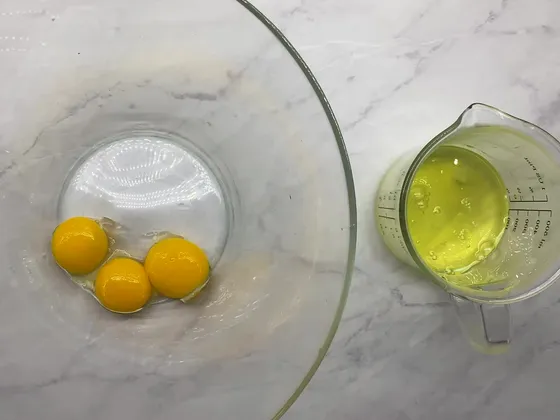 Three egg yolks in a large glass bowl next to a measuring jug of egg whites, on a marble counter