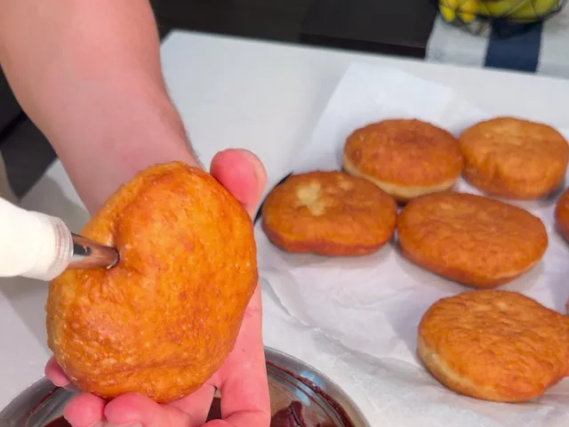 Pushing a piping tip into the side of a fried donut held in one hand, with more fried donuts on paper behind