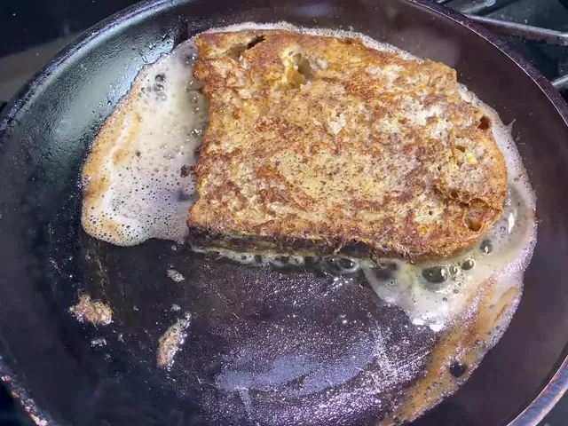 A slice of French toast frying in foaming butter in a dark frying pan, golden brown on top