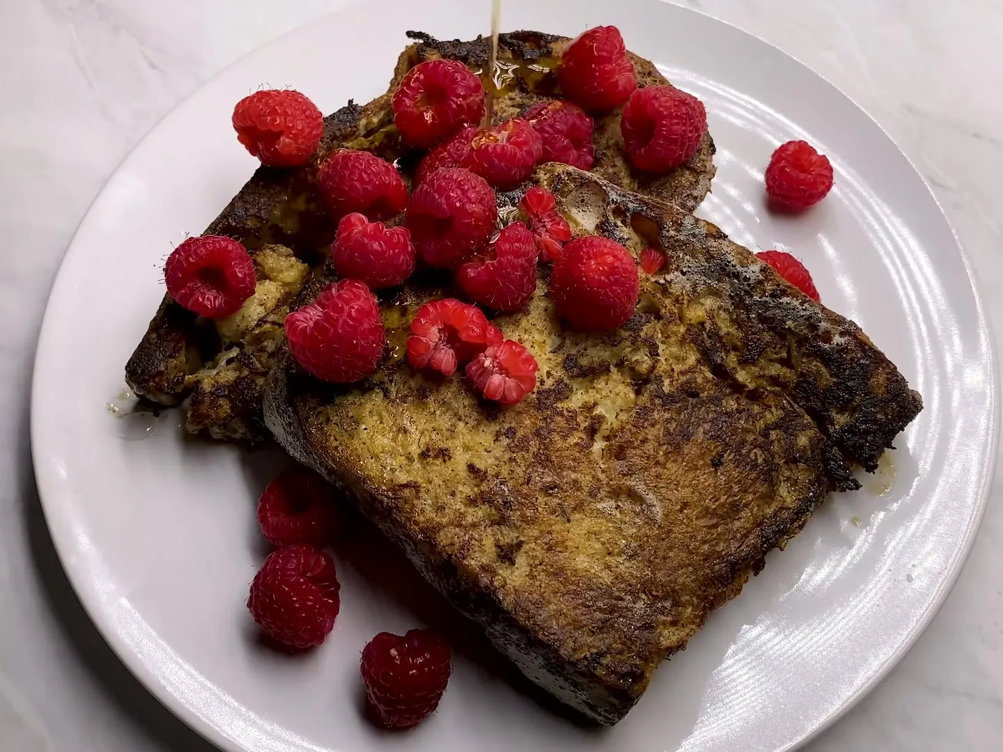 Two slices of dark golden French toast on a white plate, topped with raspberries while maple syrup is poured over them
